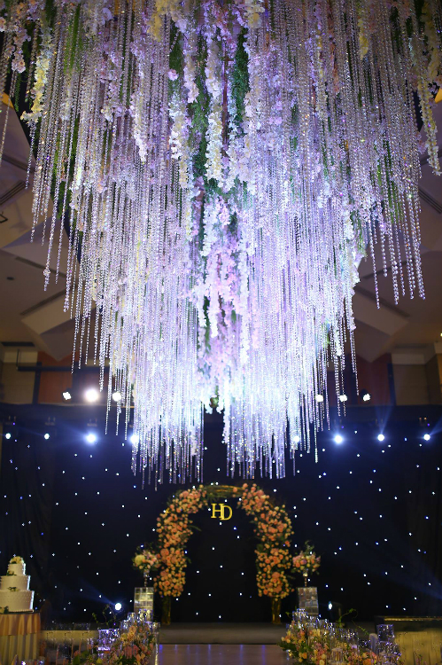 Cùng với hoa tươi, hệ thống ánh sáng cũng là điểm đặc biệt trong hôn lễ này. Một chiếc đèn chùm bằng pha lê được treo ở khu vực sân khấu, xem kẽ là những dải hoa tử đằng mềm mại.